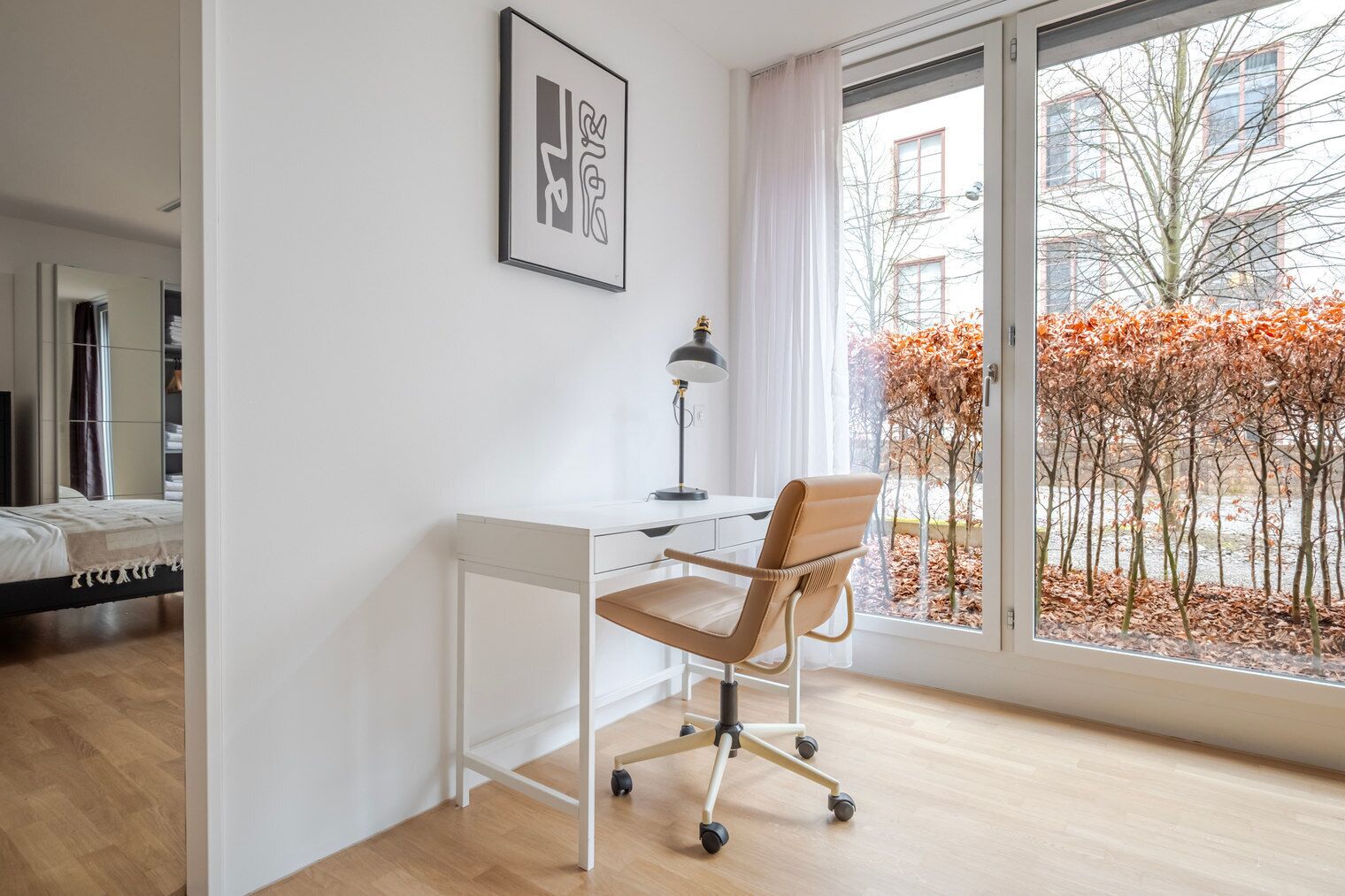 A modern bedroom with a bed, a desk with a chair, a window with a view of trees and buildings, and a framed artwork on the wall.