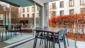 Un espace de restauration en plein air doté d’un mur de verre, d’une table avec des chaises et d’une vue sur un bâtiment et le feuillage automnal.
