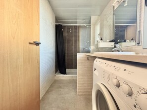 Een kleine badkamer met een wasmachine, een wastafel met spiegel en een douche met gordijn.