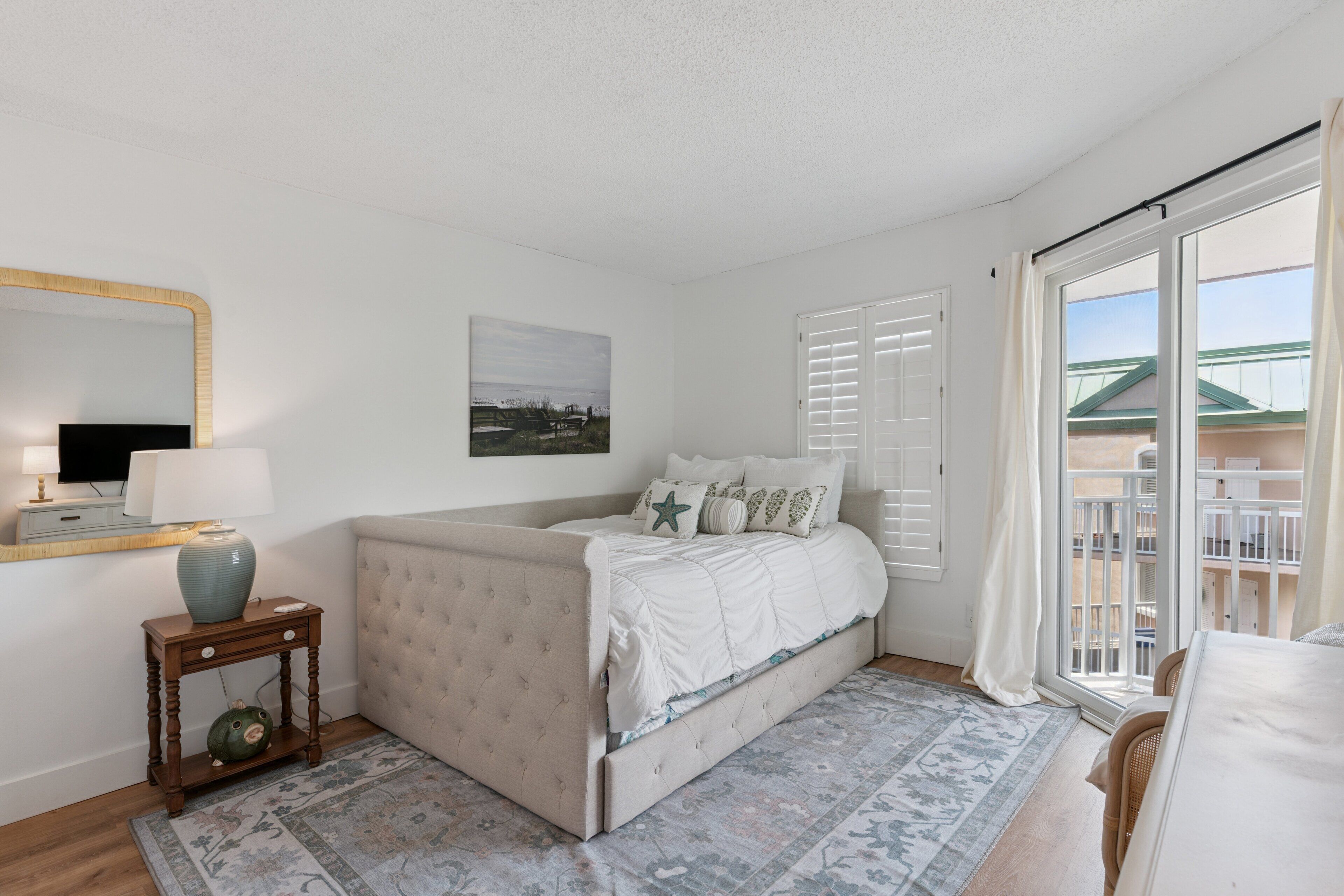 A bedroom with a bed, a bedside table with a lamp, a window with shutters, and a view of a building outside.