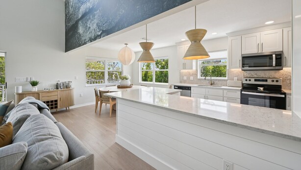 A modern kitchen with a large island, pendant lighting, and a view of greenery outside. - Salt + Sol | Heated Pool |Canal-Front | Golf Cart | Dock (Anna Maria)