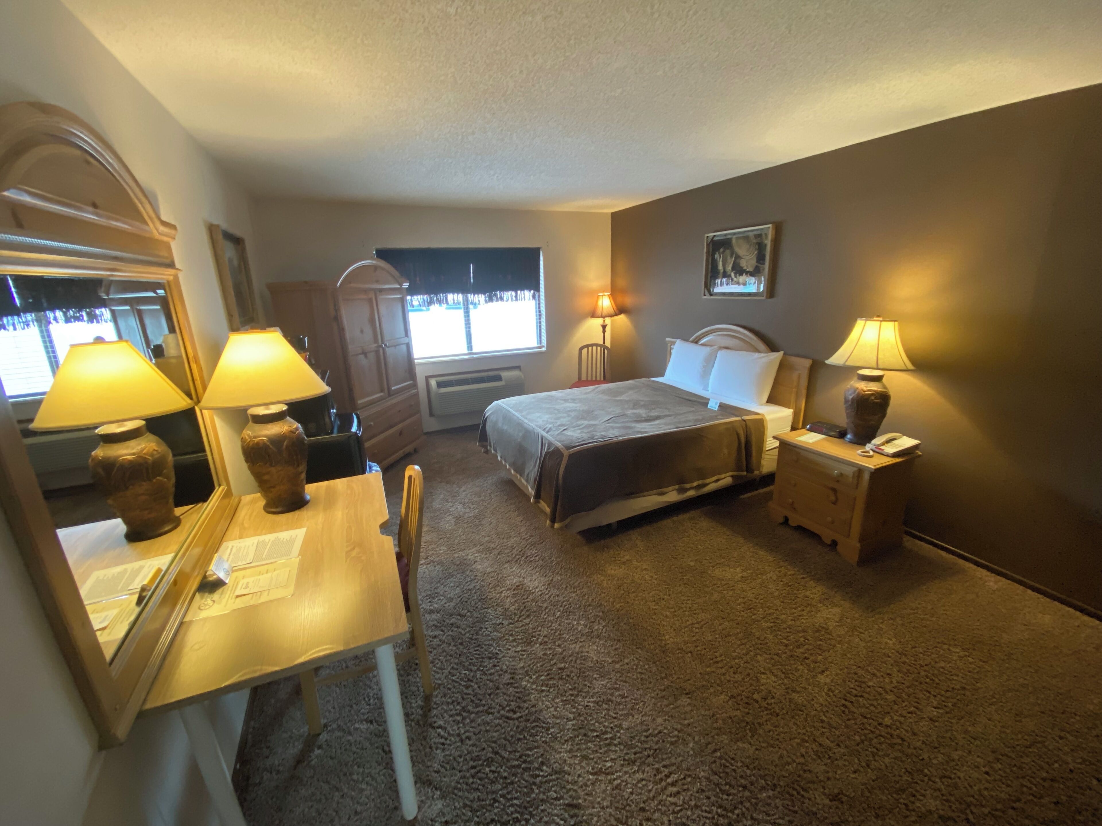 A hotel room with a bed, bedside lamps, a desk with a chair, a dresser, and a window with blinds.