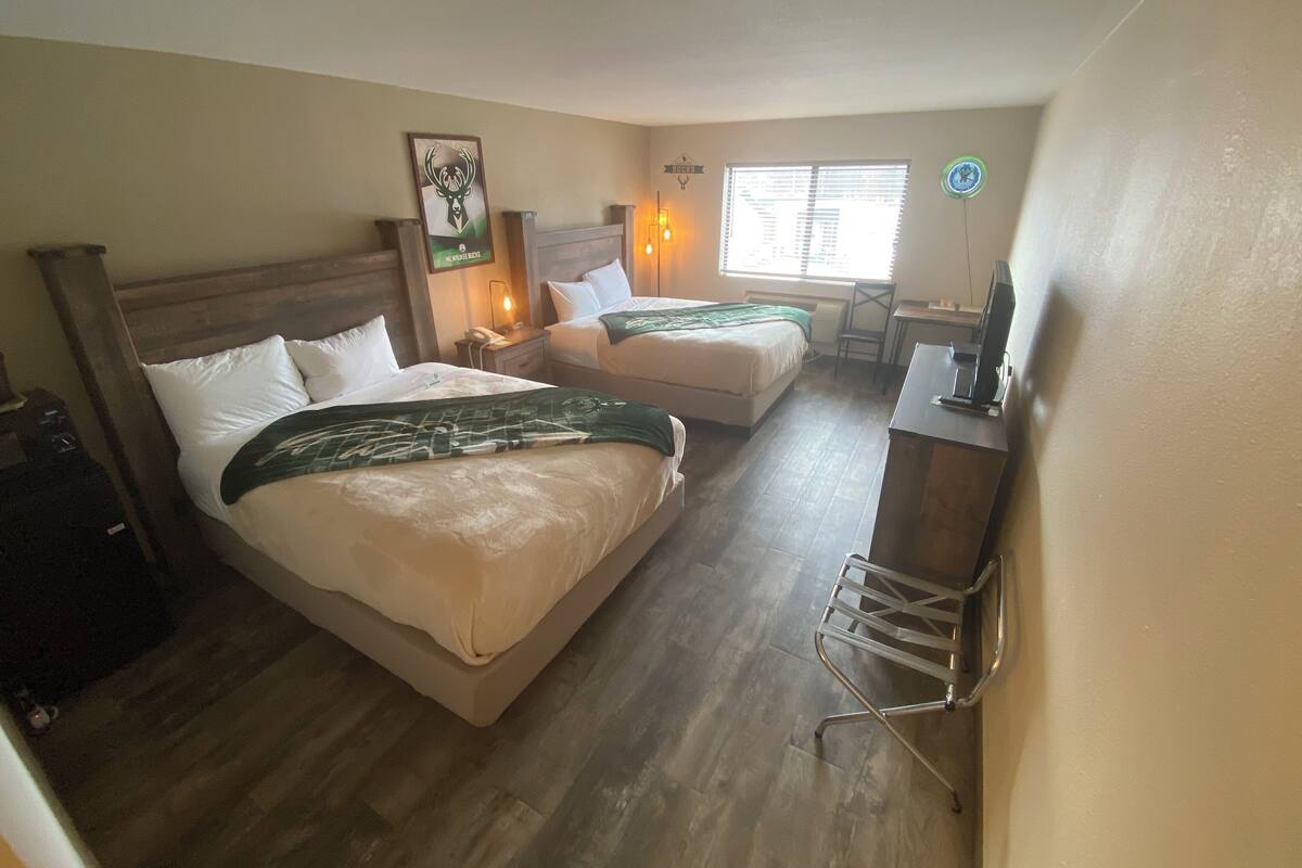 Habitación de hotel con dos camas, un escritorio, una silla, un reloj y una ventana con persianas.