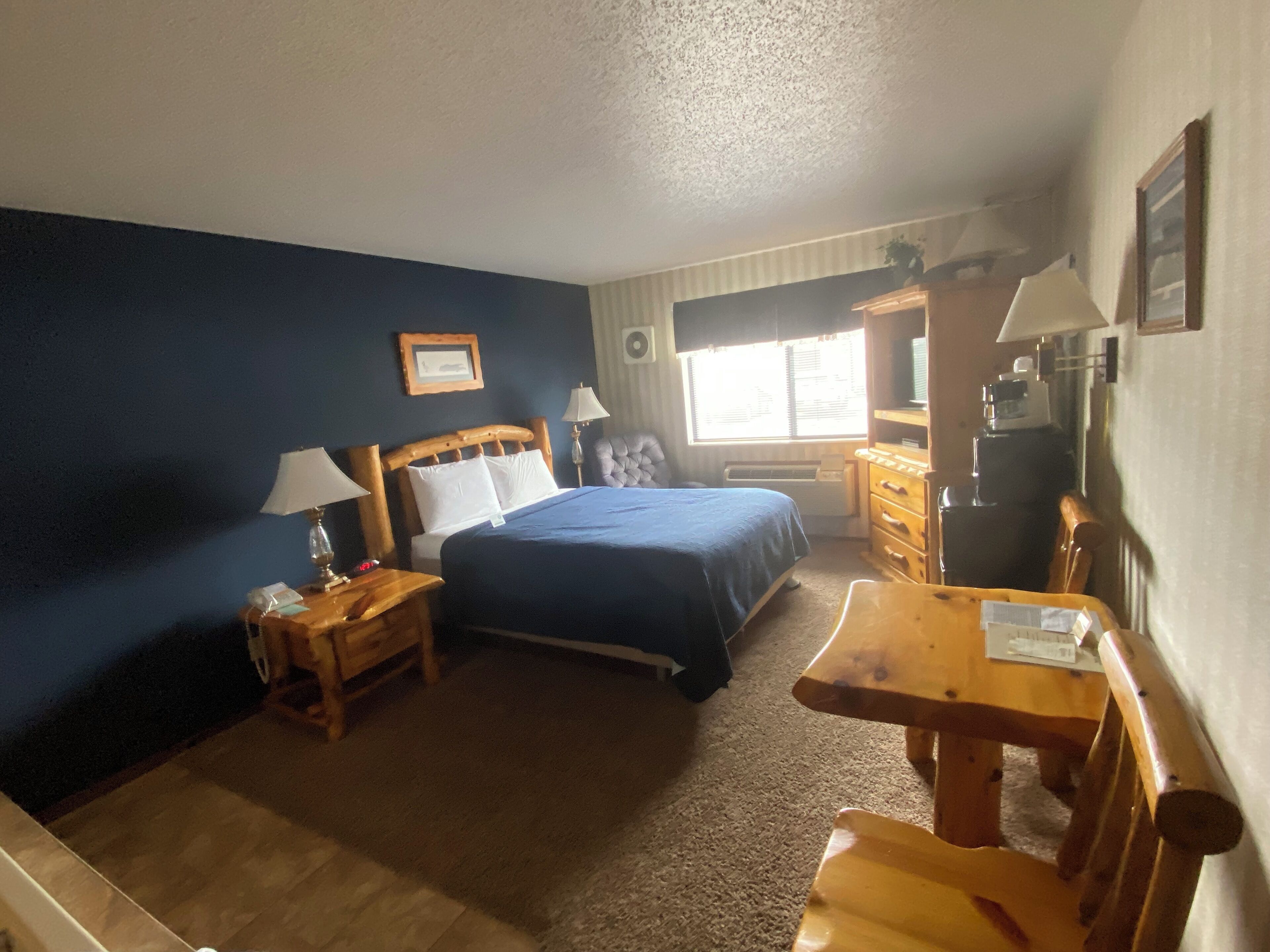 A hotel room with a bed, a desk, a chair, a nightstand, a lamp, a picture frame, a window, and a radiator.