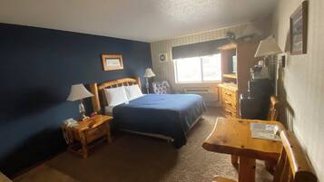 A hotel room with a bed, a desk, a chair, a nightstand, a lamp, a picture frame, a window, and a radiator.