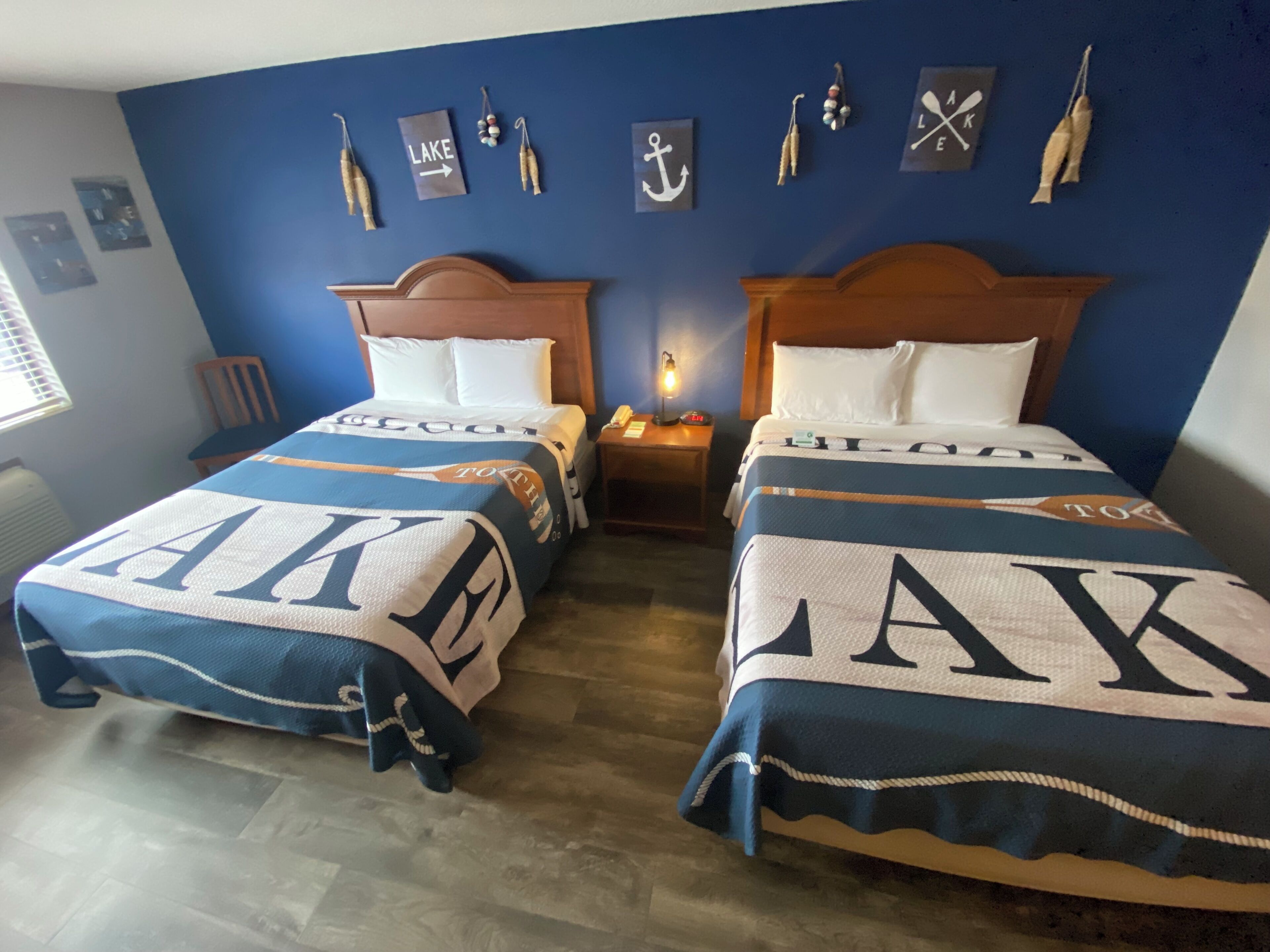 A hotel room with two beds, each with a 'LAKE' themed bedspread. There are wall decorations including an anchor and a sign that says 'LAKE'.
