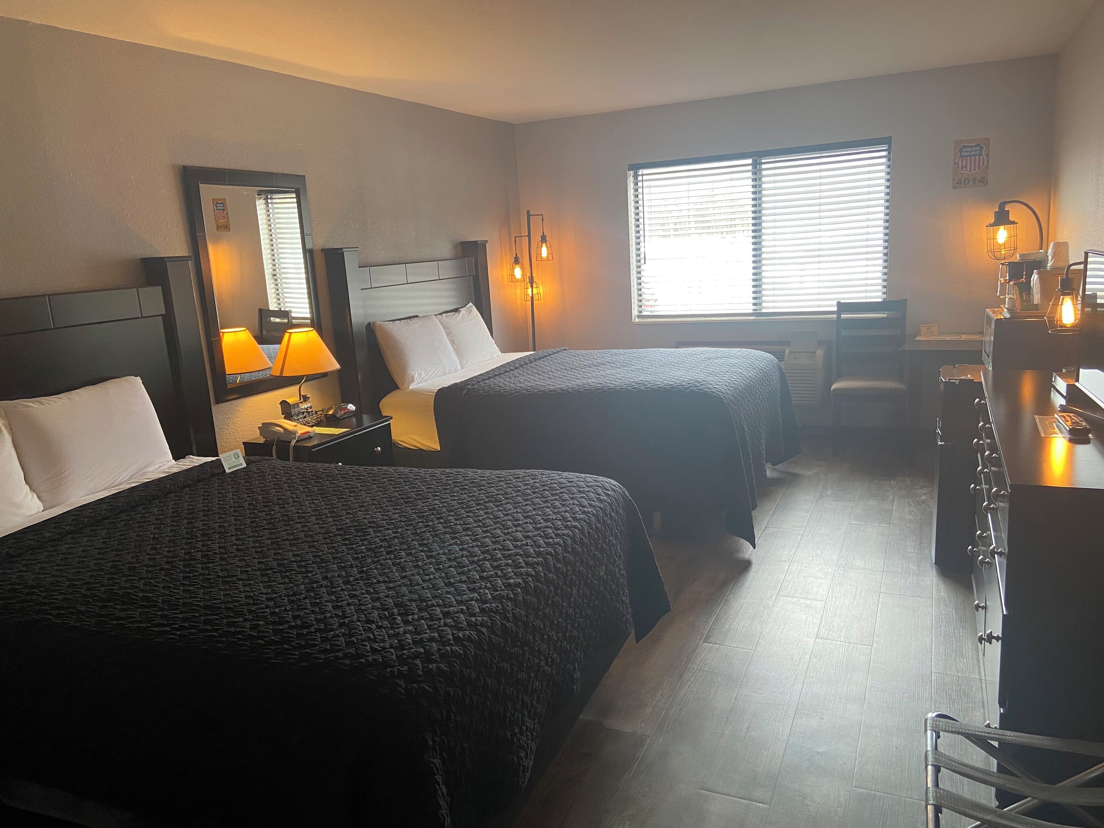 Ein Hotelzimmer mit zwei Betten, einem Schreibtisch, einem Stuhl, einer Lampe und einem Fenster mit Jalousien.