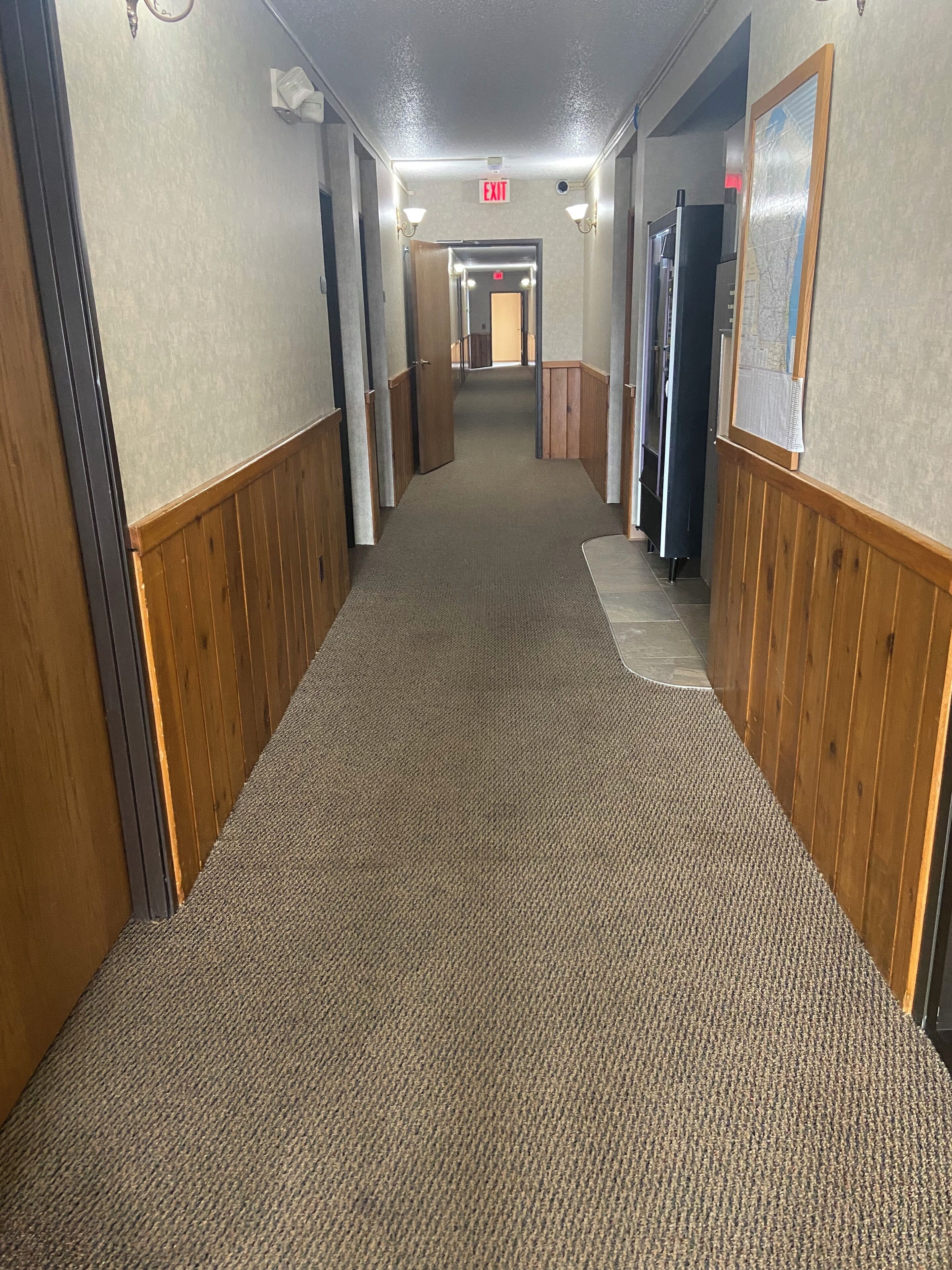 A carpeted hallway with wooden paneling on the walls, an exit sign, and a door at the end.