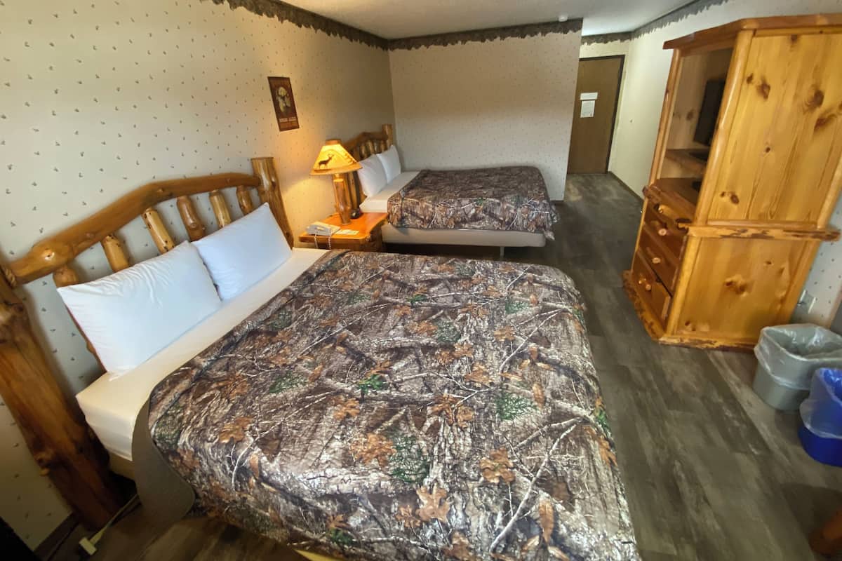 Habitación con dos camas, cabeceros de madera, una mesita de noche con lámpara, un aparador de madera y un papel tapiz estampado.