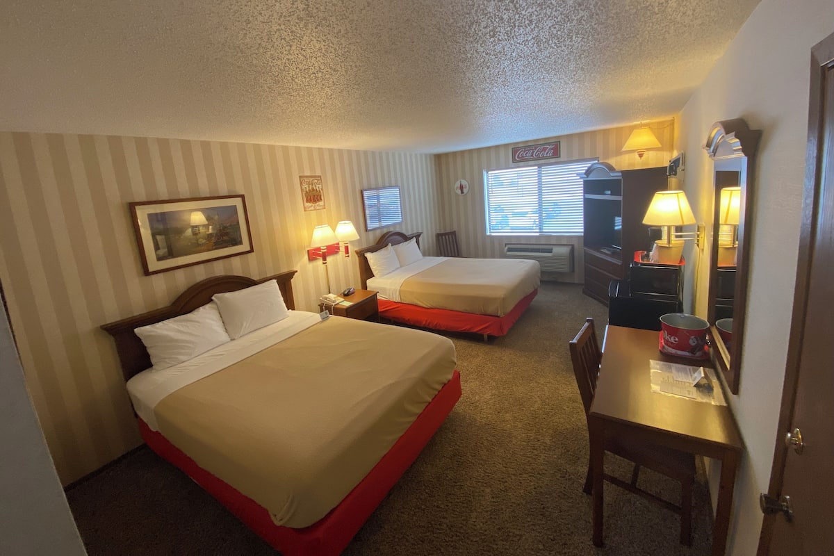 Habitación de hotel con dos camas, un escritorio con silla, cómoda, espejo y ventana con persianas.