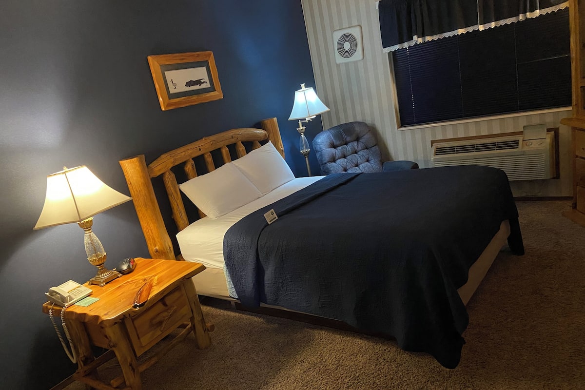 Un dormitorio con una cama de madera, una mesita de noche con lámpara, una silla y un aire acondicionado instalado en la pared.