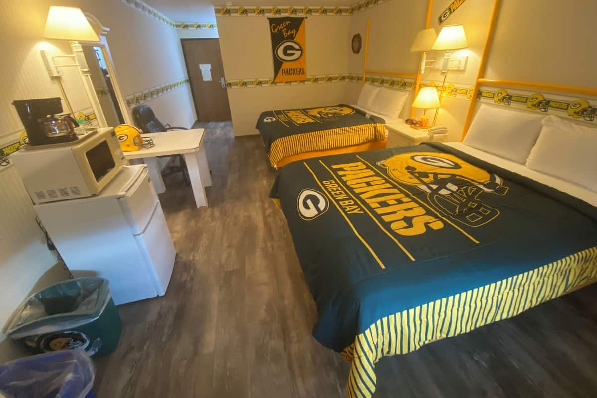 Habitación de hotel con escritorio, dos camas con ropa de cama de los Green Bay Packers, microondas, refrigerador y un cesto de basura.