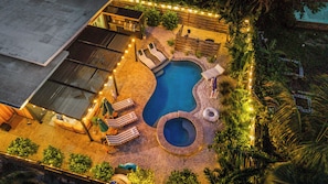 Una piscina con sillones de descanso y un jacuzzi, rodeada por una terraza de madera y luces colgantes.