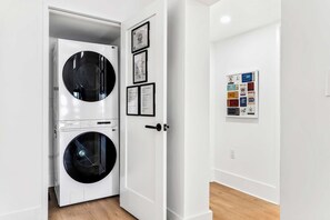 Un cuarto de lavandería con lavadora y secadora de carga frontal, una puerta con picaporte y una pared con diplomas enmarcados y un tablón de anuncios con varios avisos.