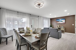 A modern living room with a dining area, featuring a large TV, wooden paneling, and a view of the outdoors. - Charming 2BR Home w heated pool in Clearwater, perfect for 5 guests near the beach! (Clearwater)