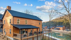 Een houten chalet met terras, uitkijkend op een meer, omringd door bomen en andere chalets.