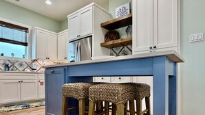 A kitchen with white cabinets, a blue island, and two woven stools. - Private Pool, Shared Pool, Boardwalk to the Beach (Port Aransas)