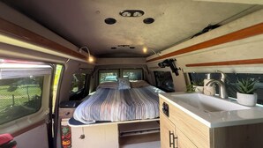 A compact RV interior with a bed, kitchenette, and a small bathroom. - World Famous  Big Wave Camper (Wailuku)
