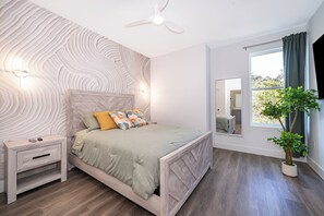 Ein Schlafzimmer mit Bett, Nachttisch, Nachttischlampe, Deckenventilator, Fenster mit Vorhängen und einer Topfpflanze.