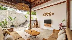 A modern living room with a stone wall, wooden furniture, a TV, and decorative items. - Villa Ivory 8Min Beach Pecatu byOrivista (Pecatu)