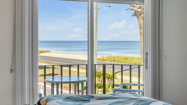 A room with a bed, a balcony with a view of the beach and ocean, and a small round table.