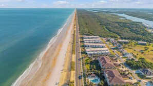 An aerial view of a coastal area with a long beachfront, a road parallel to the beach, and a cluster of buildings with a uniform architectural style.