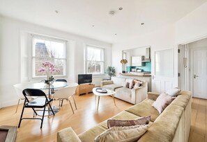 Ein geräumiges Wohnzimmer mit beigefarbener Couch, einem Esstisch mit Stühlen, einem Fernseher und großen Fenstern mit Blick auf Gebäude.