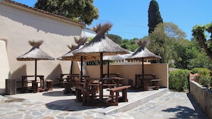 Patio esterno con pavimento in pietra, tavoli di legno e ombrelloni in paglia.