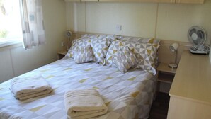 Un letto rifatto con cura, coperta con motivo geometrico, due cuscini e asciugamani piegati. Un comodino con lampada e ventilatore. Nella stanza, mobili in legno e una finestra con tende.