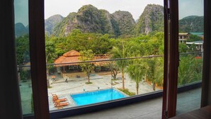 Blick aus einem Zimmer mit großem Fenster auf einen Pool, ein Reetdachgebäude und eine üppige grüne Landschaft mit Bergen.