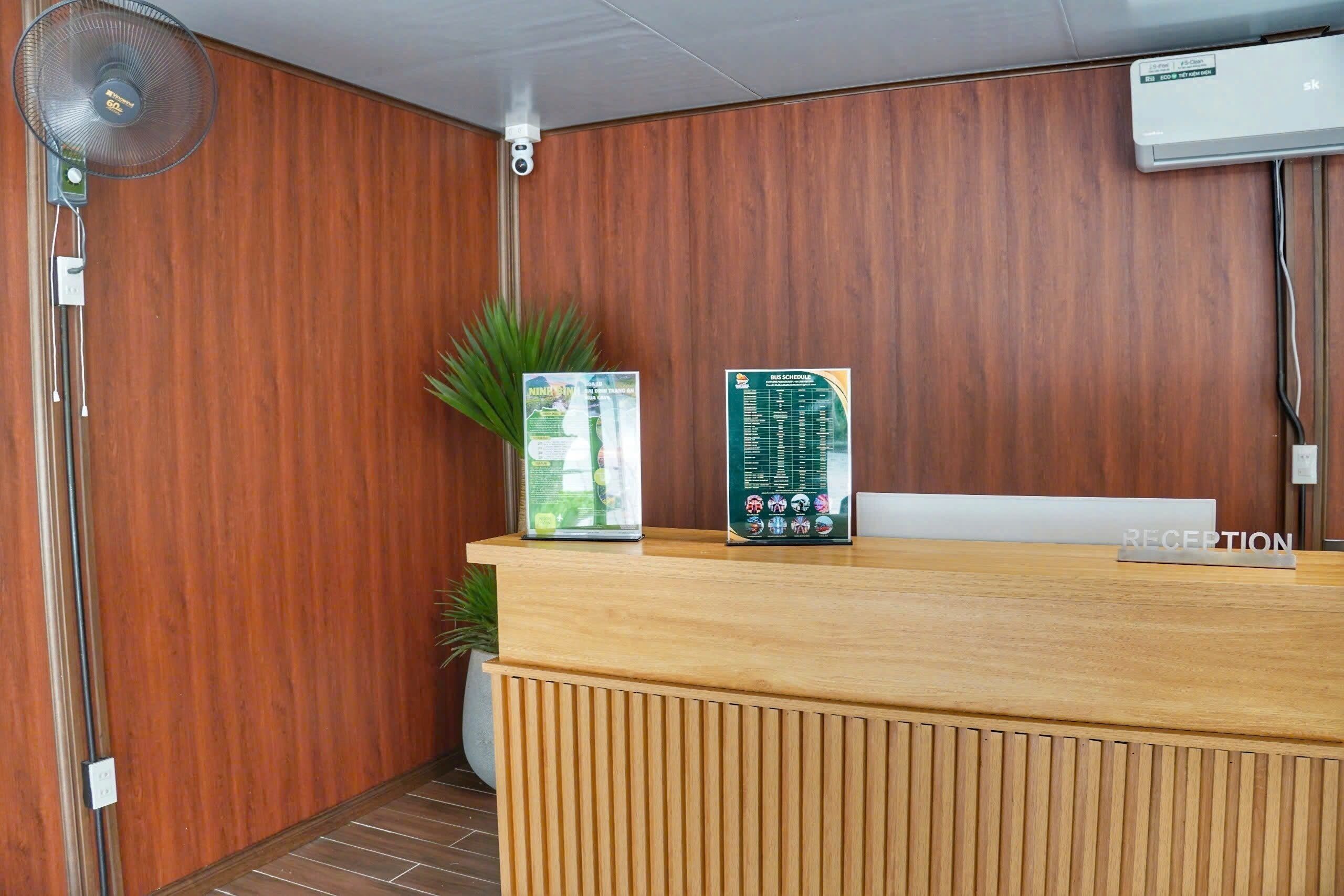 A reception desk with a wooden finish, a wall-mounted air conditioner, a floor fan, and a potted plant.