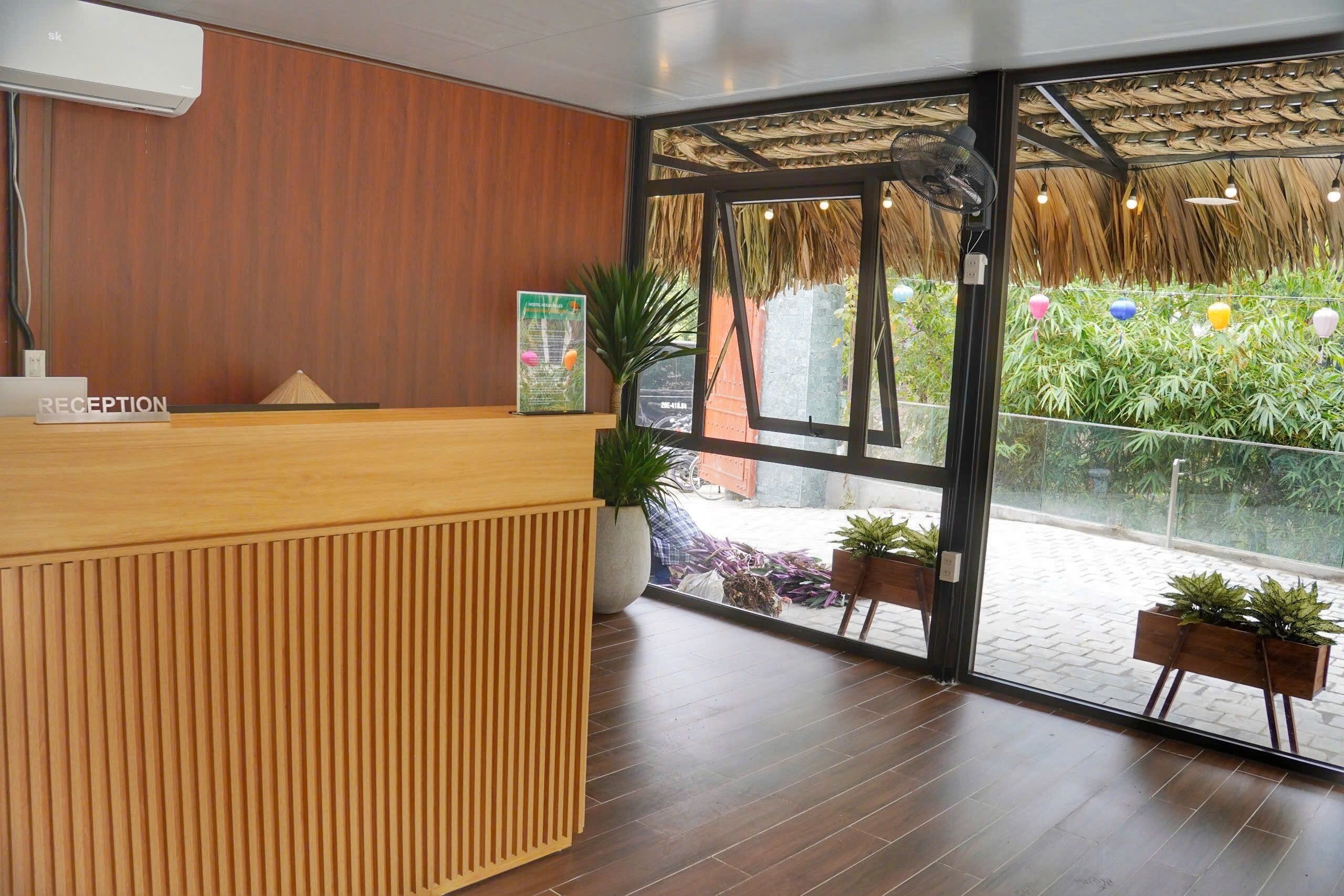 Un mostrador de recepción con detalles de madera, una planta grande en maceta y una puerta de cristal que da a un espacio exterior con techo de paja y vegetación.