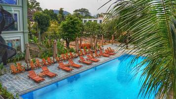 A pool area with sun loungers and umbrellas, surrounded by greenery and a modern building with a spiral staircase.