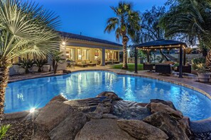 A swimming pool with a rock feature, a covered seating area, and a house with large windows.