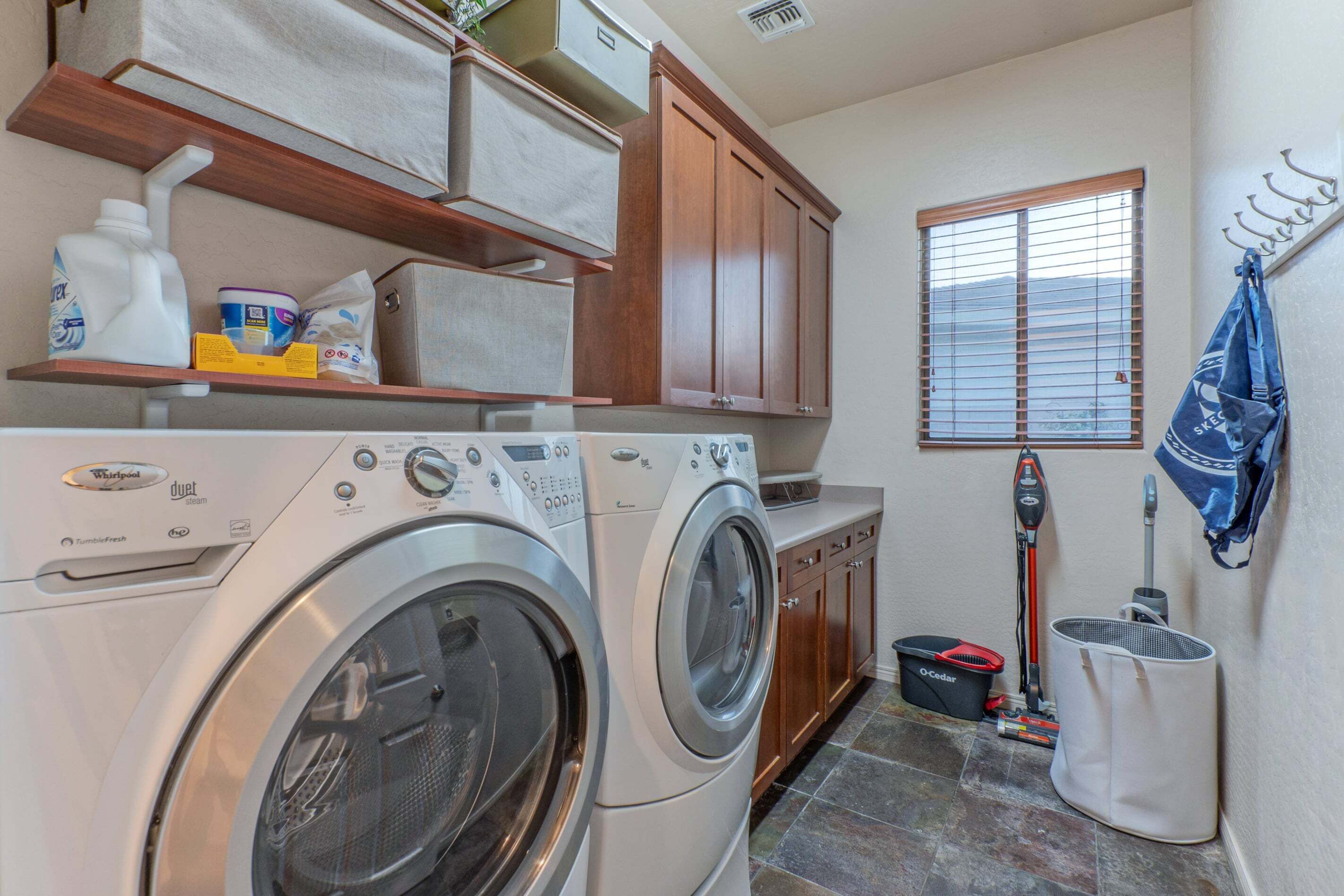 A laundry room with two front-loading washers, wooden cabinets, a window with blinds, and a mop on the floor.