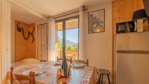 A dining area with a table set for two, a bed with a wooden headboard, a refrigerator, and a view of mountains through a window. - Cham Sud Iris361 Near Aiguille du Midi | Balcony Views, Chamonix, France (Chamonix)