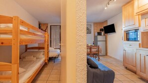 A room with bunk beds, a sofa, a dining table, and a kitchen area with cabinets and a TV. - Cham Sud Iris361 Near Aiguille du Midi | Balcony Views, Chamonix, France (Chamonix)