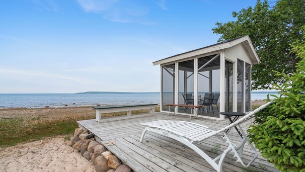 En träbrygga vid stranden med ett avskärmat sällskapsrum med glasväggar, en bänk och ett bord.