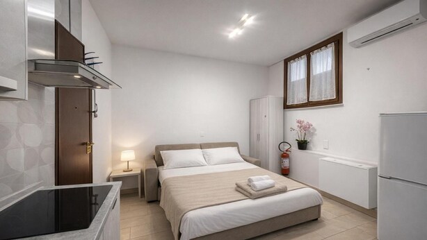 Ein kompaktes Hotelzimmer mit Bett, Nachttisch, Lampe und einer Küchenzeile.