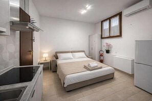 Una habitación de hotel compacta con cama, mesita de noche, lámpara y un área de kitchenette.
