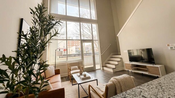 A modern living room with a large window, a sofa, a coffee table, a TV, and a staircase. - Boulevard Stays Midtown 30ft Ceilings 1BR 01 (Nashville)