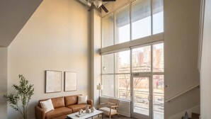 A spacious living room with a high ceiling, a brown leather sofa, a coffee table, and a large window offering a view of the outside. - Boulevard Stays Midtown 30ft Ceilings 1BR 01 (Nashville)