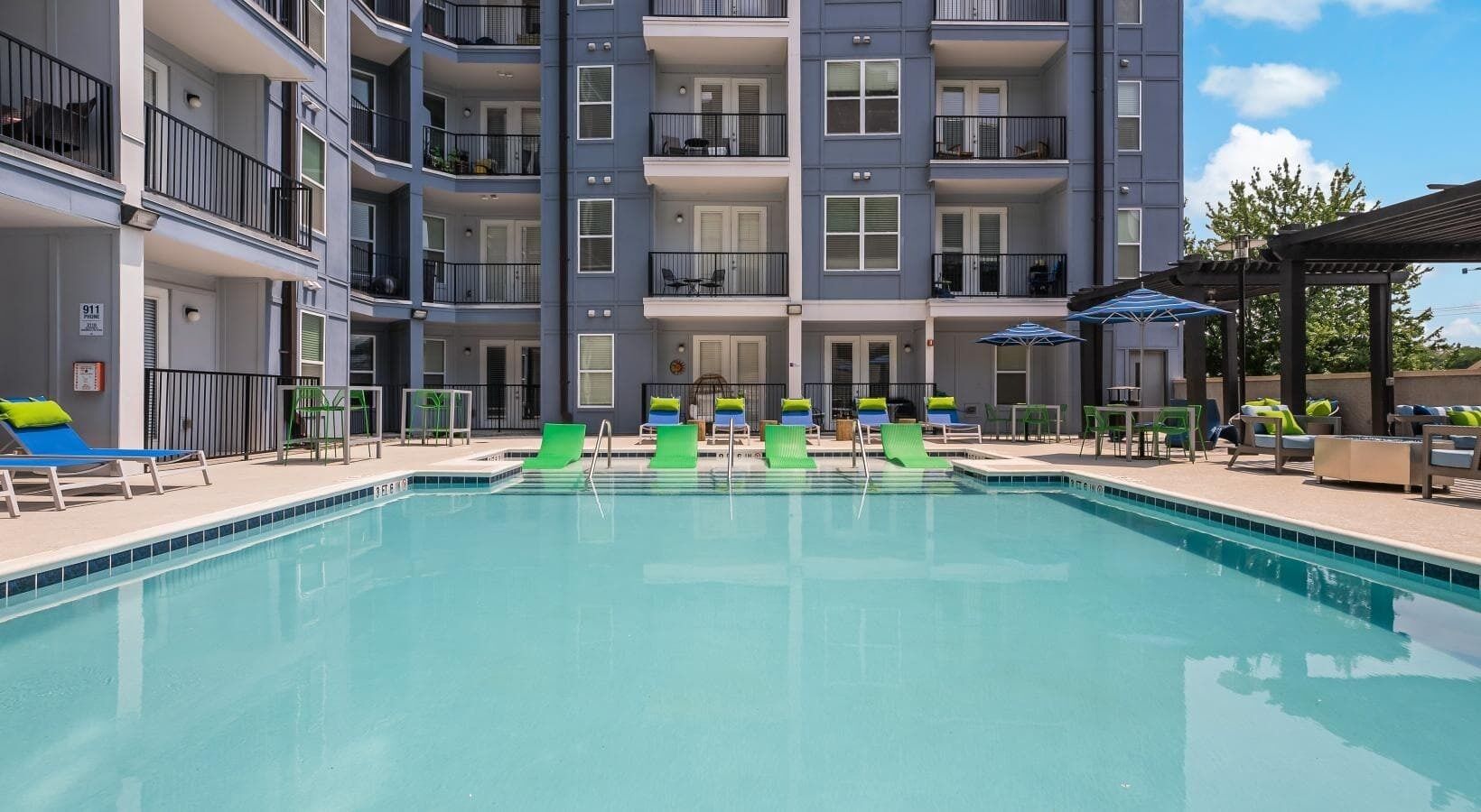 Une piscine entourée de chaises longues et de parasols, elle-même bordée d’un immeuble à plusieurs étages doté de balcons.