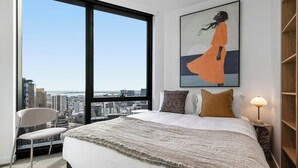 Un dormitorio con un ventanal grande, una cama con una colcha estampada, un almohadón, una lámpara de noche y vistas a la ciudad.