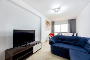 Una sala de estar moderna con televisor de pantalla plana, un sofá de color oscuro, una mesita con lámpara y una ventana con cortinas.