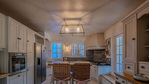 Una cocina amplia con armarios blancos, una isla central con sillas, una lámpara colgante y vistas a través de las ventanas.