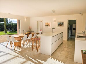 A modern kitchen with a central island, white cabinetry, and a dining area with a table and chairs. - Exceptional Air-Conditioned Villa with Pool for 10 Guests – Gulf of Saint-Tropez (GRIMAUD)