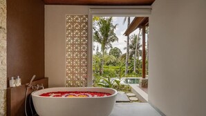 Ein Badezimmer mit einer freistehenden Badewanne, gefüllt mit bunten Badezusätzen, einer Holzbalkendecke und Blick auf einen üppigen Garten.