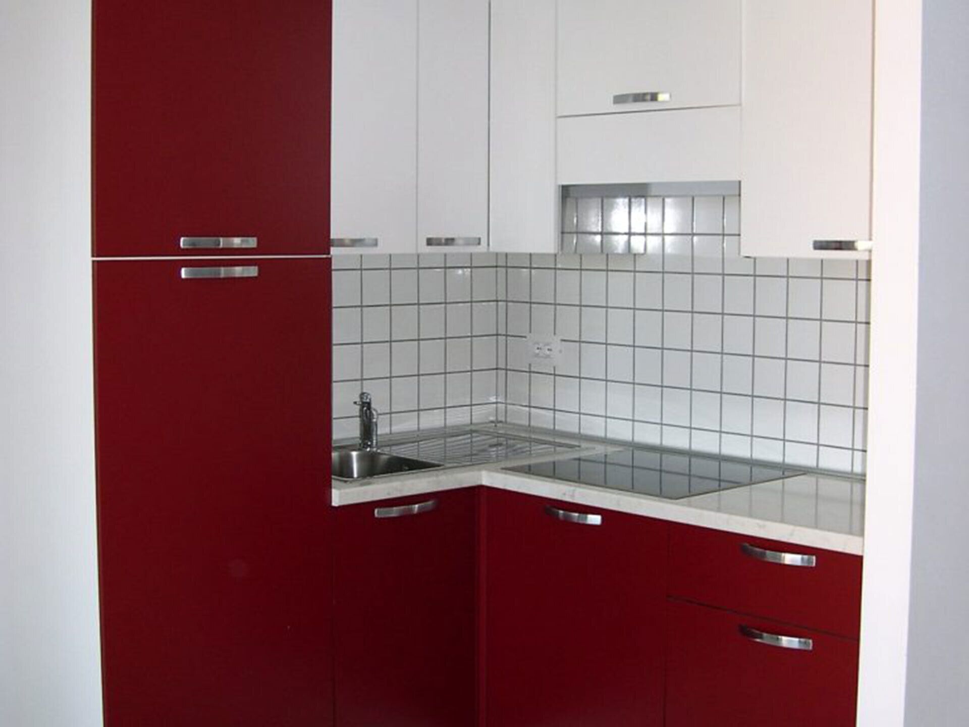 A compact kitchen with red cabinets, white countertops, and a stainless steel sink.