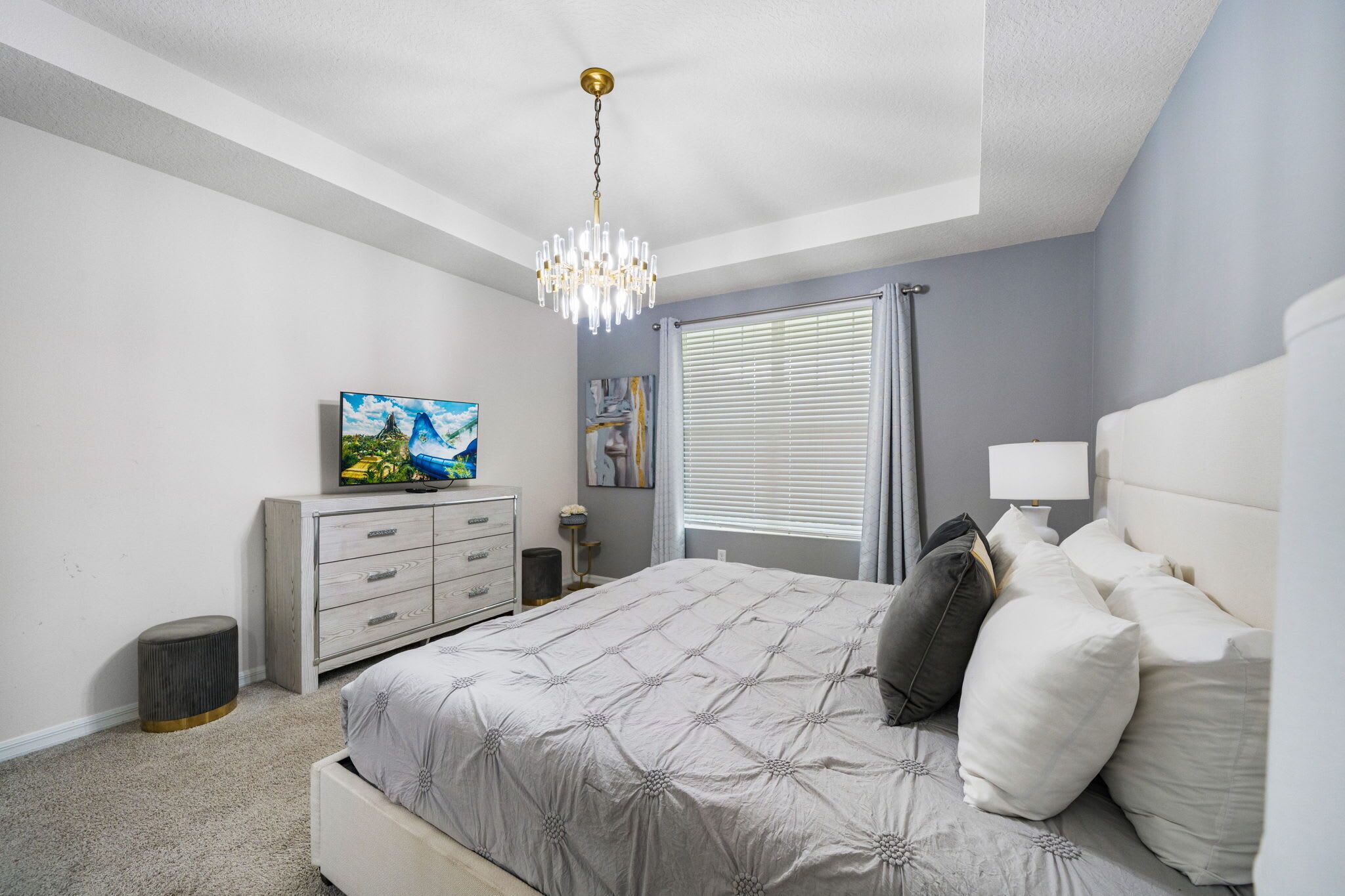 A bedroom with a bed, a dresser, a chandelier, and a window with blinds.